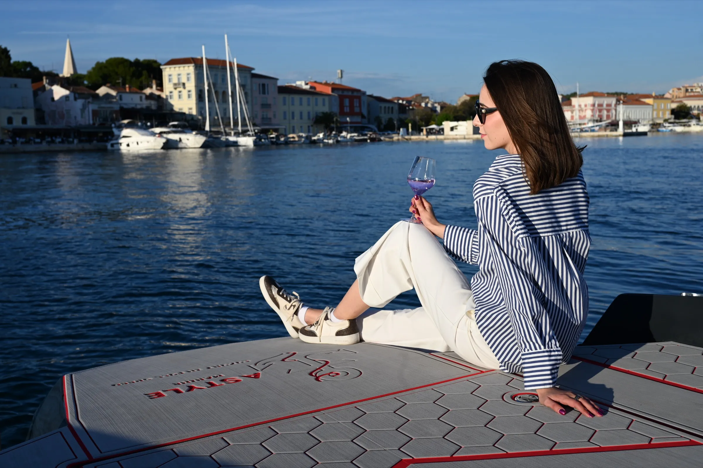 Gošća s čašom rosea sjedi na pramcu jahte i gleda preko mora prema porečkoj rivi sa zvonikom i marinom